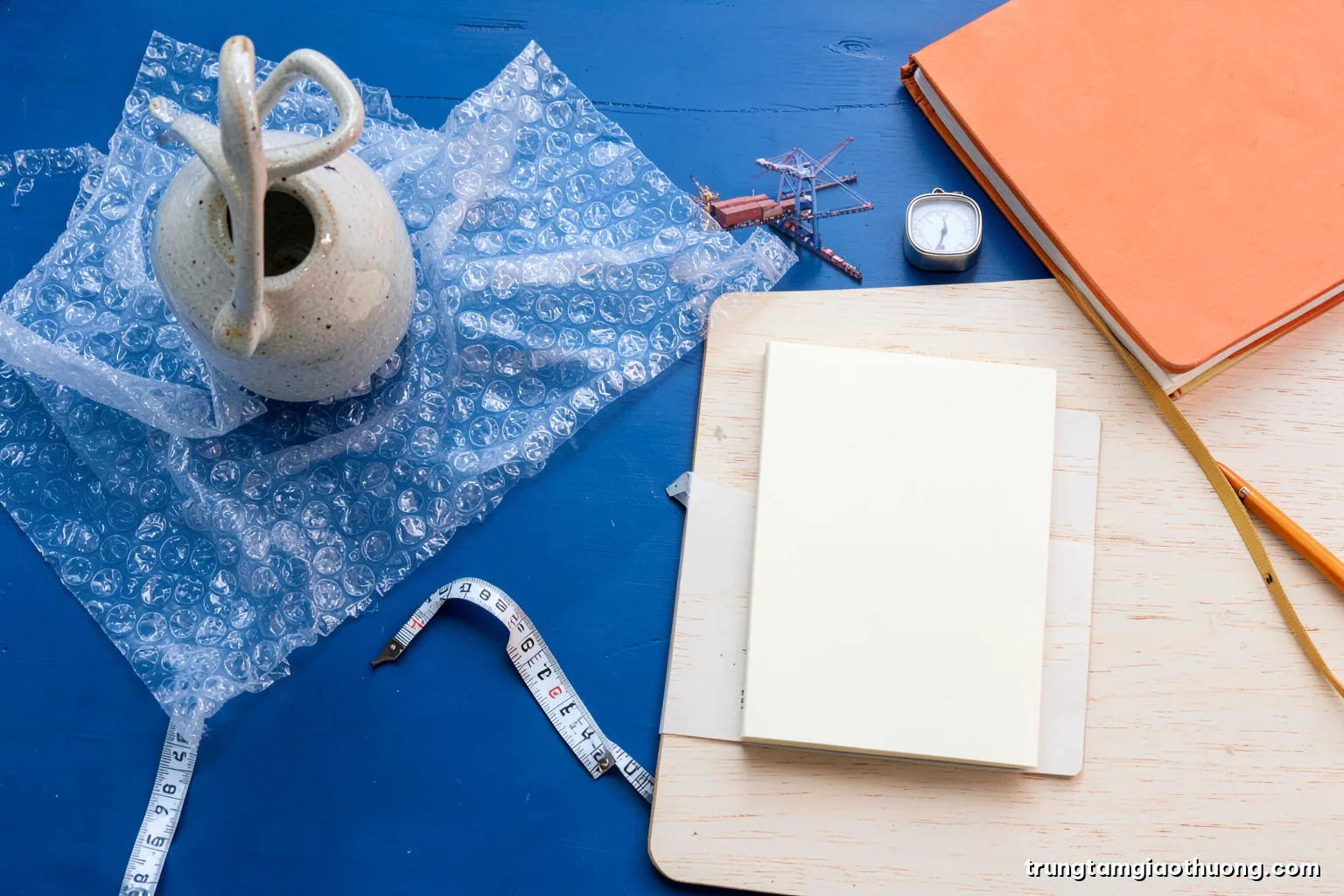 Top-down flat lay of a ceramic vase, bubble wrap, a measuring tape, a small scale and a blank notebook on a pale wooden table, crisp diffused light, terracotta and cream palette