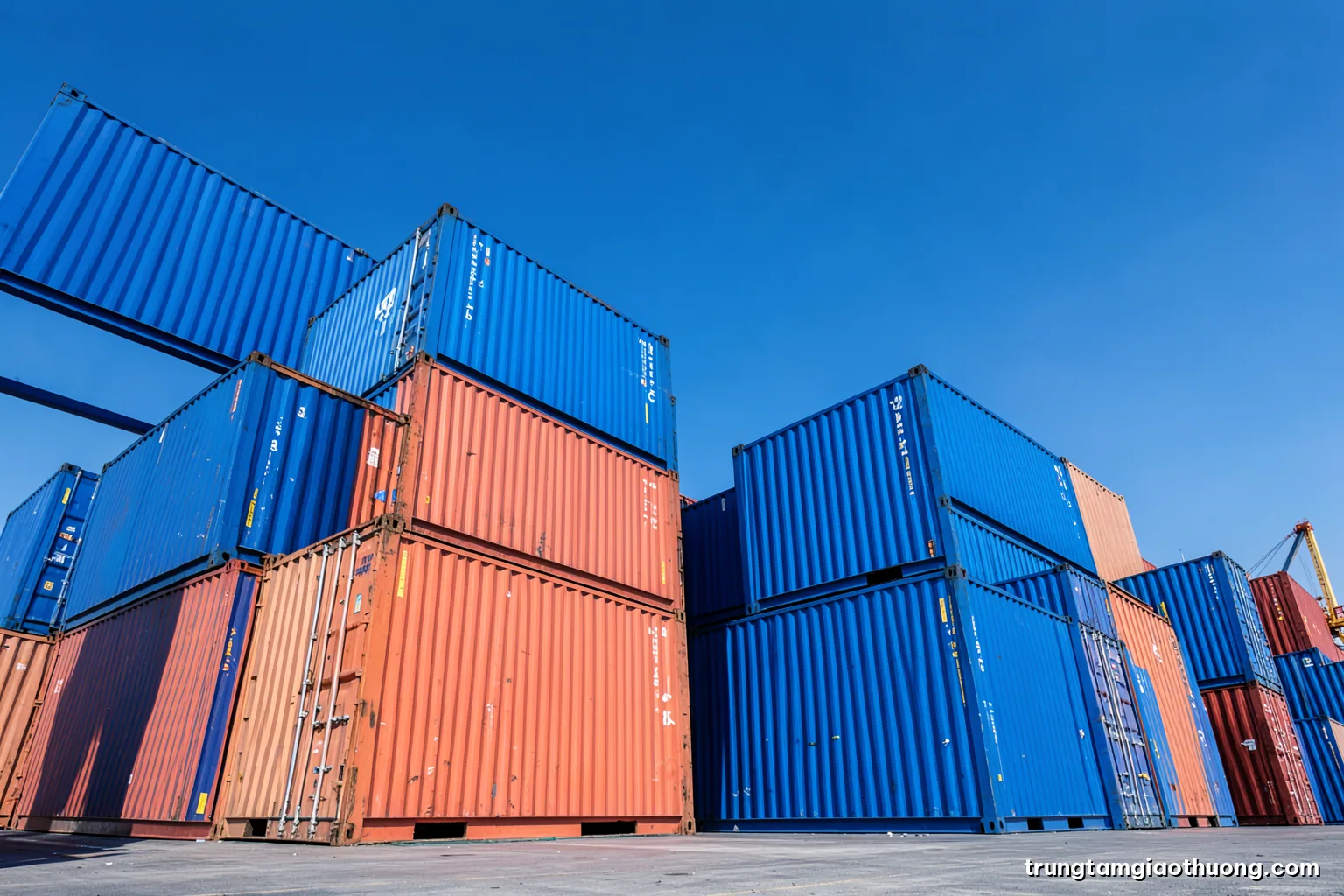 Low angle shot of stacked shipping containers in two contrasting colours against a clear sky at a Vietnamese terminal, crisp midday light, cobalt and coral palette, minimalist composition