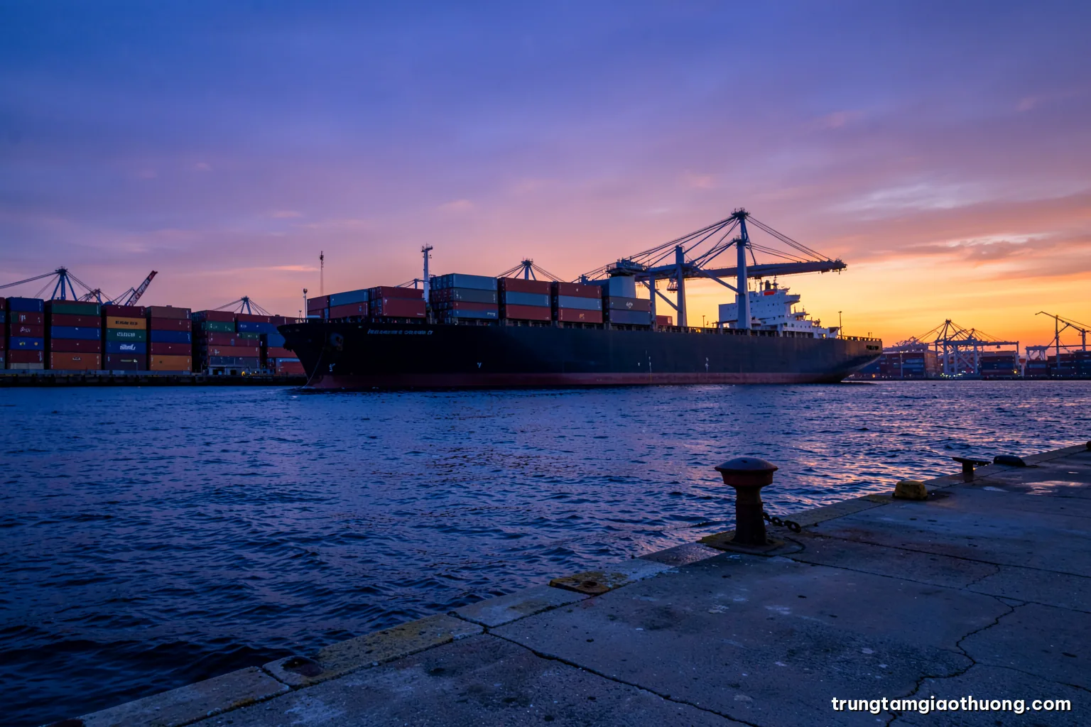 Silhouette of a large container ship leaving harbour at sunset viewed from a quiet concrete quay with mooring bollards in foreground, purple and amber sky, wide angle